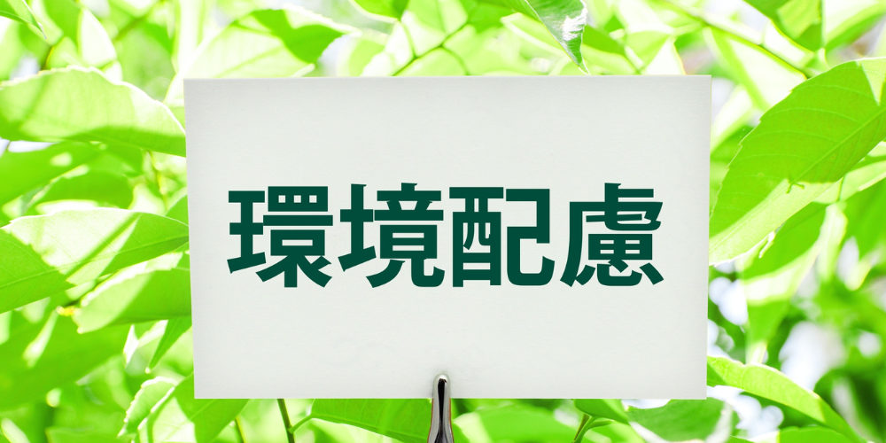 緑色の葉が茂る背景の前に白いボードが立てかけられ、太い緑文字で ‘環境配慮’ と書かれている写真