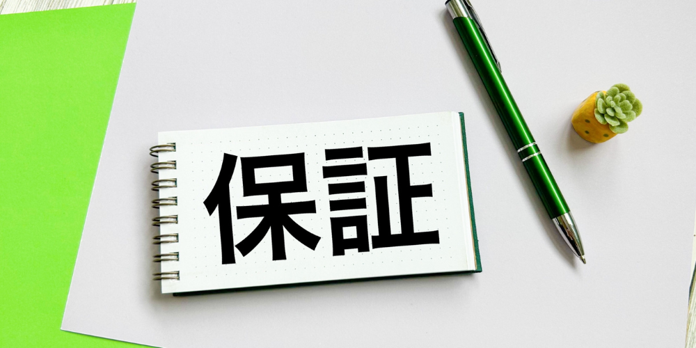 白いデスクに緑のペンと多肉植物が置かれ、その中央のノートに黒い文字で ‘保証’ と書かれている写真