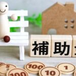 補助金の文字ブロックと木製の貨幣、家の模型や電卓が並ぶ住宅支援のイメージ