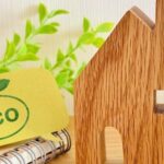 木製の家の模型と「eco」と書かれた札。観葉植物や花に囲まれたエコ・自然素材を意識した住まいのイメージ。