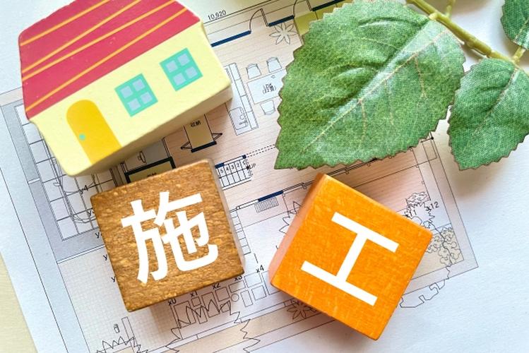 間取り図の上に置かれた家の模型と「施工」と書かれた木製ブロック、住宅建築のイメージを伝える温かみのある構成