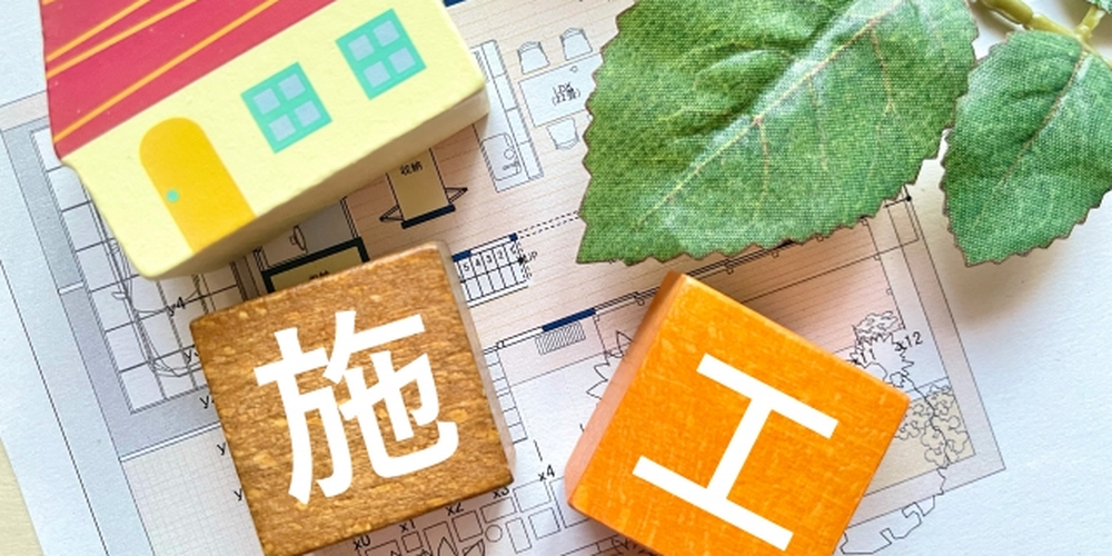 間取り図の上に置かれた家の模型と「施工」と書かれた木製ブロック、自然素材を想起させる葉の装飾