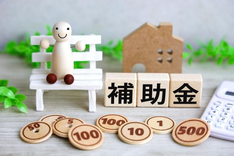 補助金の文字が書かれた木製ブロックとベンチに座る人形、家の模型や木製コインが並ぶ住宅補助金のイメージ