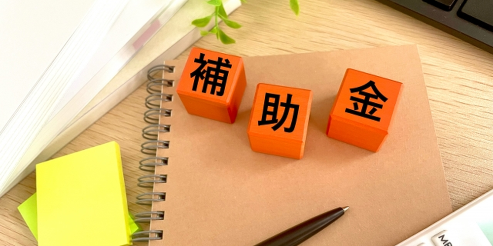 「補助金」と書かれたオレンジ色のブロックとノート、電卓が置かれたデスク上のイメージ