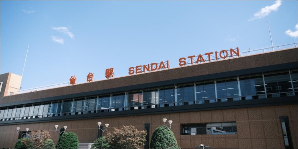 青空の下に佇む仙台駅の外観と正面の街路樹
