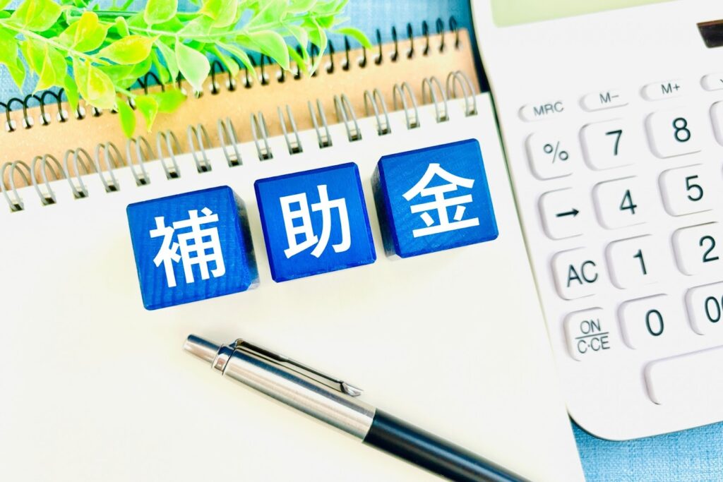 ノートブックや電卓が置かれた机の上に補助金と書かれたキューブブロックが置いてある画像